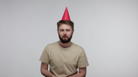 Optimistic joyful bearded guy with funny cone hat laughing and blowing confetti glitters, celebrating birthday congratulating on anniversary, festive mood. studio shot isolated on gray background