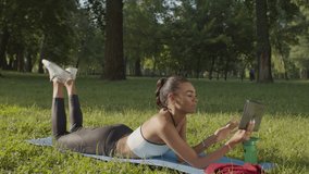 Positive attractive sporty fit african american woman lying on exercise mat on park lawn, watching fitness workout online using digital tablet during outdoors sports training in the morning.