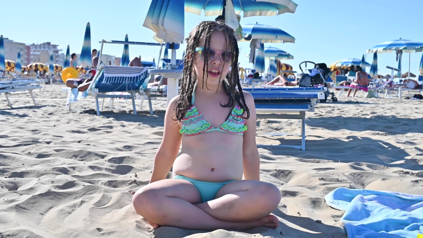 Little girl in a swimsuit on the beach plays, smiles. Child 7 years old on the equipped beach of Italy. The concept of a summer, family vacation by the sea. The person shows positive emotions. 4k