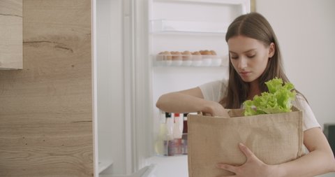 Cheerful young woman unpacking food bag into fridge. Pretty smiling housewife putting groceries in refrigerator. Online food delivery. 4k raw video footage
