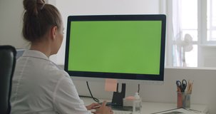 Young woman doctor work on computer with green mockup screen. Girl in uniform and lab coat clothes sitting in hospital office room using laptop. Over shoulder camera shot. 4k video footage slow motion