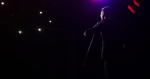 Man stand up comedian speaking jokes in micropphone in the light of the floodlights standing on stage with his back to the camera. Actor, artist on funny performance. Actively gestures with his hands.
