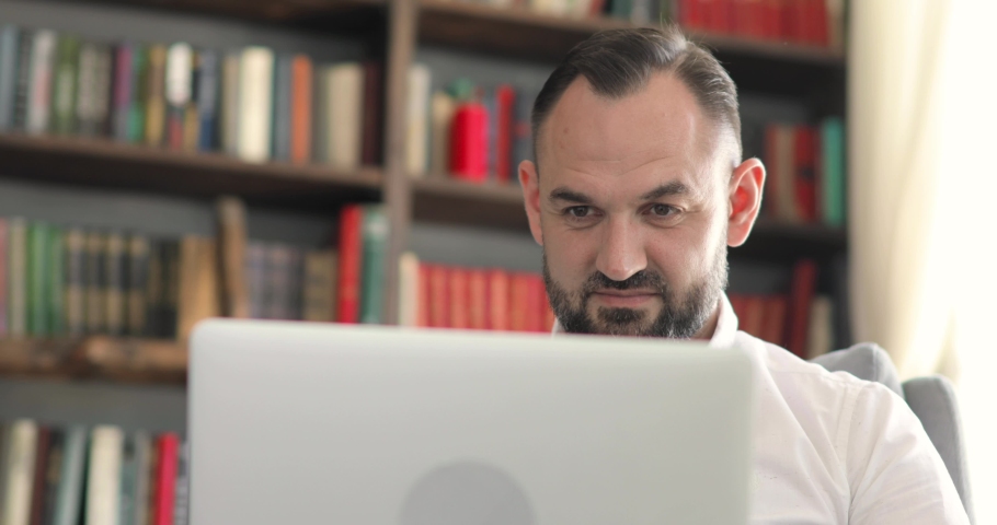 Portrait of man is talking video call sitting in office on bookshelves background. Remote work of teacher, professor or entrepreneur online at the computer, laptop in office. Remote work concept.