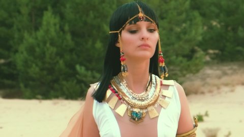 beautiful woman fashion model, queen Cleopatra walking. white luxury dress Pharaoh clothes gold accessories, tiara, necklace, jewelry. Black hair Egyptian makeup. green pine tree yellow sand desert
