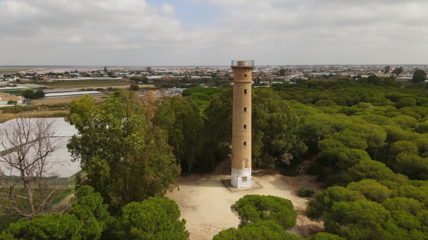 Aerial view of the slightly inland San Jeronimo lighthouse at the mouth of the Guadalquivir River in the Atlantic Ocean in western Analusia. The tower is framed by trees.