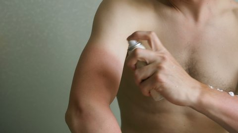 young man with bare chest applies spray remedy to cure red sunburned skin on arm sitting against light grey wall close view slow motion