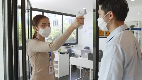 Asian businesswoman wear protective face mask using infrared thermometer check temperature of working people before enter ing office, reopen business with new normal and social distancing concept