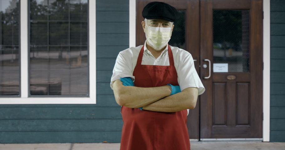 The owner of a small bakery or butcher shop stands in front of his store wearing a mask due to COVID19 mandates.