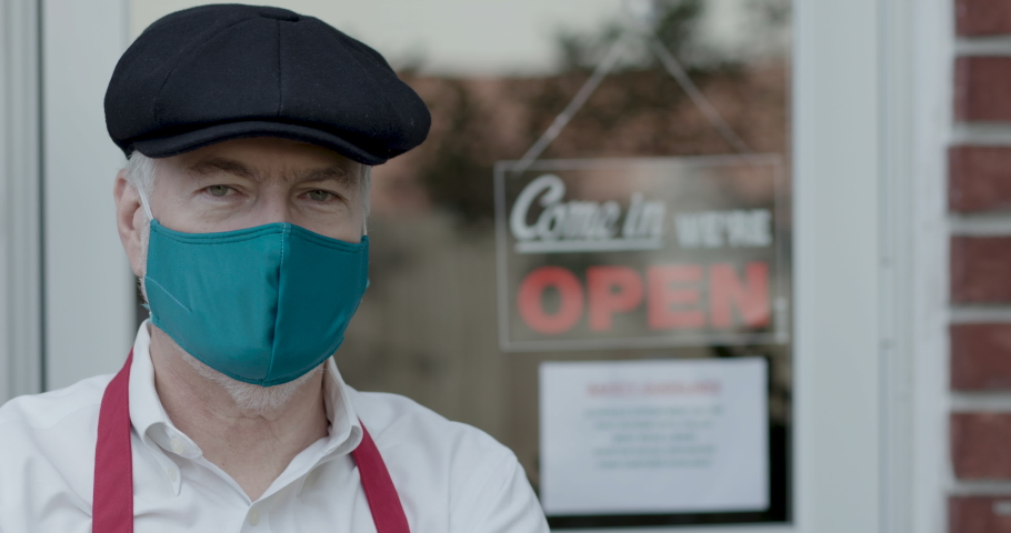 Small business owner stands in front of his store which is open for business wearing a face covering or mask due to COVID19 mandates.
