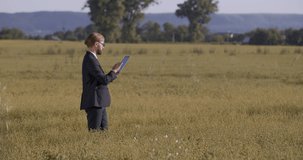 Mature owner of agricultural business dressed in black suit, taking photos of green field on digital tablet. Competent farmer in eyeglasses using modern device at work outdoors.