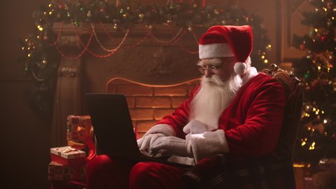 An elderly man in a Santa Claus costume is working at a laptop on the background of a Christmas tree