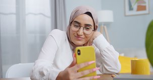 Close up of young muslim woman wearing hijab and glasses having video call using camera on yellow smartphone while sitting at cozy stylish modern home office. Young asian student speaking with family.