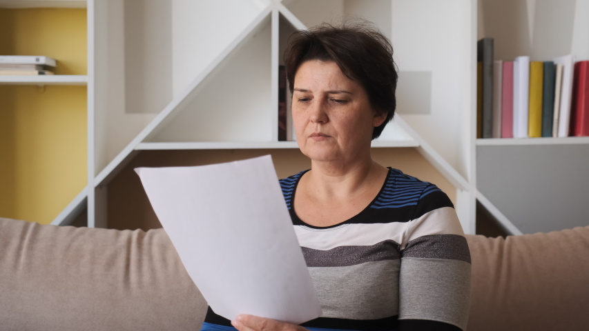 Sad woman sitting on couch at home reads received bad news holds documents paper letter feels desperate about financial problems. expulsion concept Royalty-Free Stock Footage #1055844278