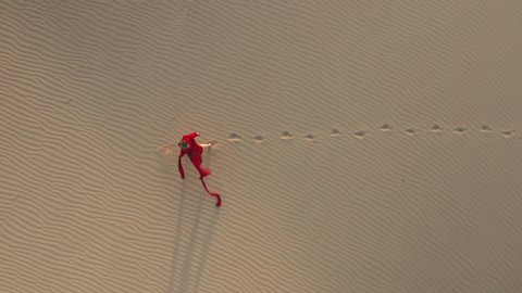 Nature landscape shot. A lonely traveler in fashion red fashionable fluttering dress is walking by the beige sand dunes. 4K aerial slow motion video of a beautiful female model in the desert at sunset