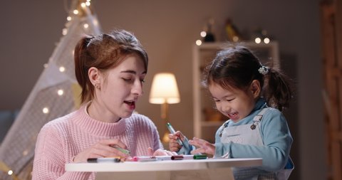Cute little asian girl playing with her mother at home, drawing together with markers. Babysitter is teaching the kid how to draw - childhood, parenthood concept 4k footage