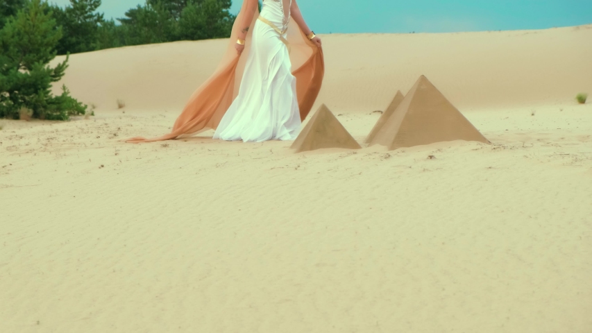 mysterious silhouette beautiful female legs. Woman walks on sandy desert Egypt. White long vintage dress waving in slow motion, orange silk cape. Backdrop decoration Egyptian pyramid, green trees