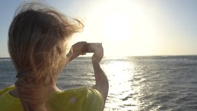 Woman standing alone on sunny windy morning tropical beach and taking selfies to place pictures at social media resources. Person using digital camera of mobile smartphone.