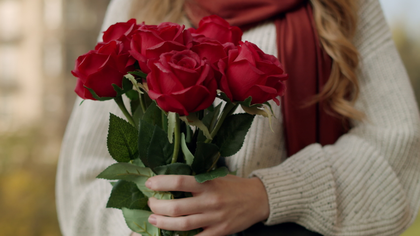 Closeup woman hands bringing flowers to face outdoors. Portrait of sweet hipster girl smelling bouquet of flowers on city street. Happy lady enjoying bunch of roses outside.
