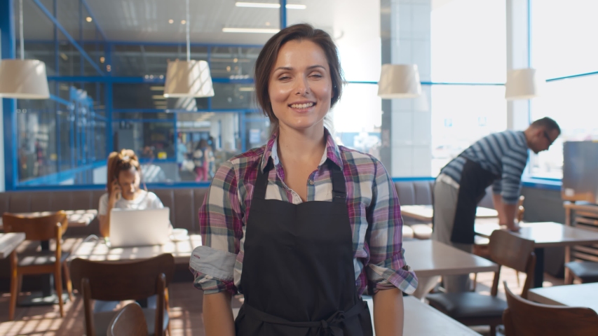 Portrait of cheerful waitress in apron smiling at camera with hands crossed standing in cafe. Smiling young staff or small cafe business owner entrepreneur looking at camera, posing in cafeteria Royalty-Free Stock Footage #1055869412