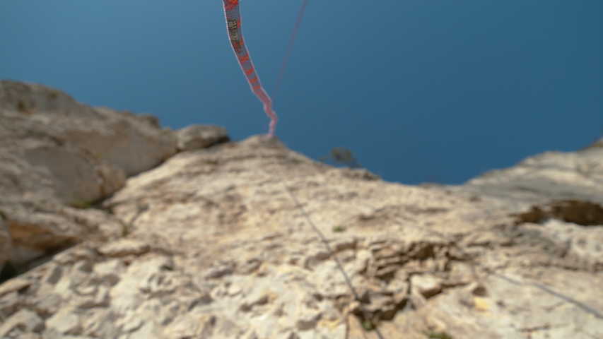 SLOW MOTION, BOTTOM UP, DOF: Thin blue and red ropes hang from the top of a towering cliff on a sunny summer day. Safety ropes get fitted after the lead climber reaches the summit of a rocky wall.