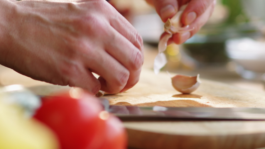 Close-up shot of hands of male cook crushing garlic clove with knife and then trimming the root end and peeling it above wooden cutting board