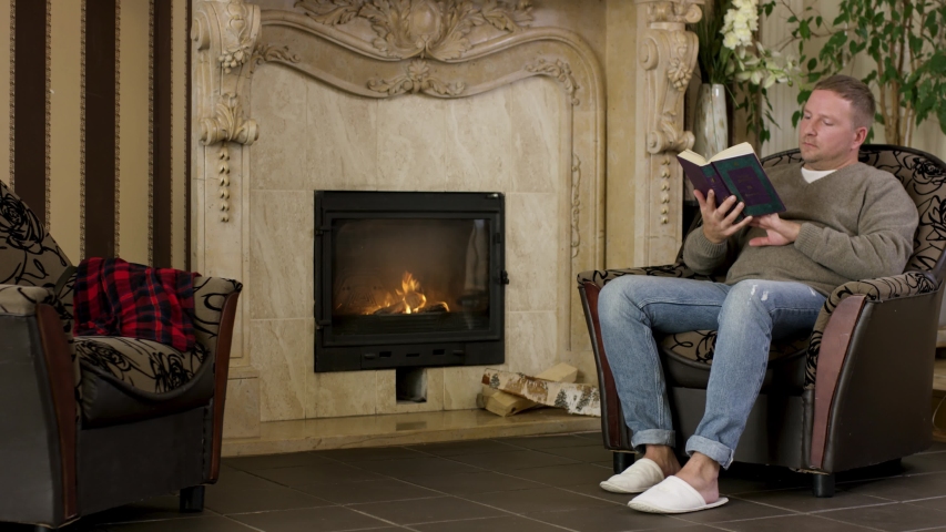 Young man sitting in a chair at home, reading a book in front of fireplace. Video. Male wearing pullover and jeans sitting at home by the fire and reading.