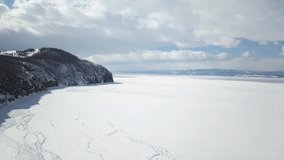Stunning natural landscape of Siberian wild nature. Clip. Aerial view of ice and snow surface of Lake Baikal in the background Olkhon island in a winter cloudy day.