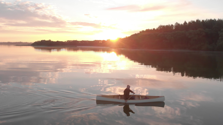 AERIAL Mid 50s Caucasian male canoeing on a traditional wooden boat on a large lake early in the morning. 4K UHD sunset shot