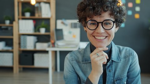 Slow motion portrait of attractive young businesswoman in glasses smiling in creative office looking at camera. Happy people, occupation and business concept.
