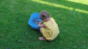 A little girl with pigtails in a yellow summer dress is playing with toys in the garden on the grass.