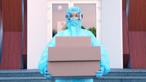 Courier, in protective uniform, mask, goggles, holds in hands some cardboard boxes, on background of entrance door to hospital. Cargo delivery service during coronavirus outbreak.