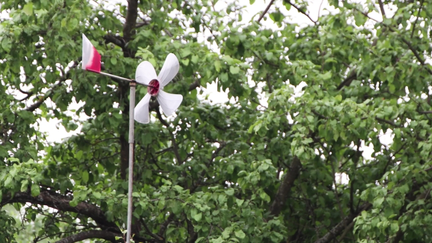 A white weather vane with a red tail without wind does not spin. On the background of green dense trees. Summer warm day. Soft light. UHD.