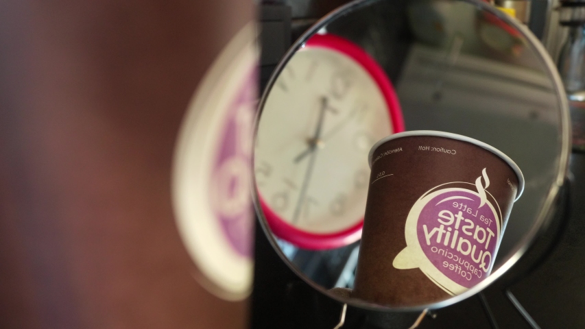 Reflected view of water being poured into disposable cup. Clocks are at background.