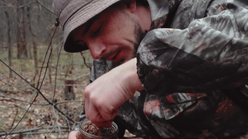 Hunter forest ranger in camouflage suit eats canned mushrooms from jar. Man with mustache in panama hat.