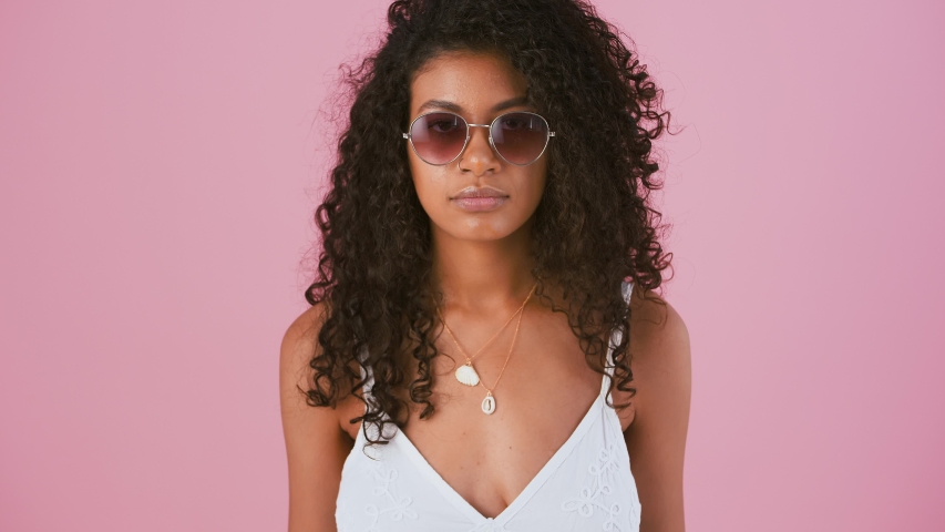 Dark-skinned woman in sunglasses, white dress. She is smiling, pointing at you by forefingers while posing on pink studio background. Close up Royalty-Free Stock Footage #1055923631