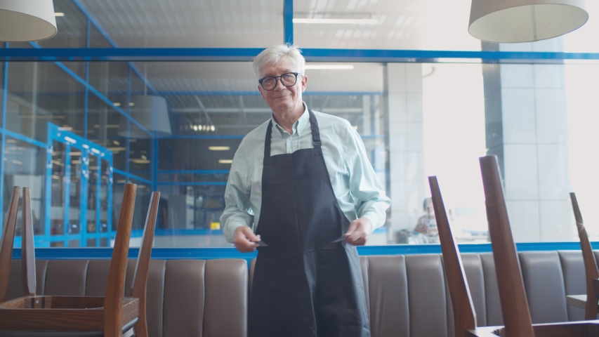 Mature man small business owner arranging apron walking in closed cafe. Senior male restaurant manager preparing for opening new place or reopening after quarantine | Shutterstock HD Video #1055924990
