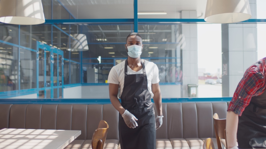 Portrait of african waiter in apron safety mask and gloves walking in cafe with colleague wiping table with cloth on background. Small business during quarantine concept | Shutterstock HD Video #1055925002