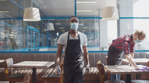 Portrait of african waiter in apron safety mask and gloves walking in cafe with colleague wiping table with cloth on background. Small business during quarantine concept