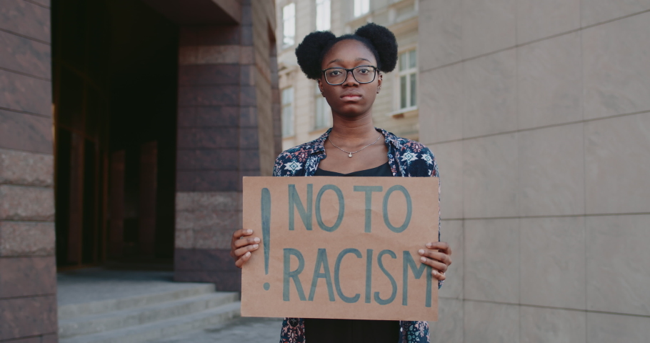 Portrait of african american girl looking to camera while holding placard no to racism. Woman with nose ring protesting against racial inequality at city street. Concept of human rights. Royalty-Free Stock Footage #1055925179