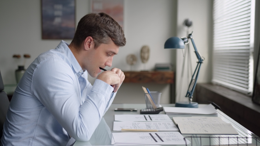 young serious business man with glasses sits at table in office, works with papers. has difficulties, gets upset because of failure and problems, shakes head. side shot Royalty-Free Stock Footage #1055925359