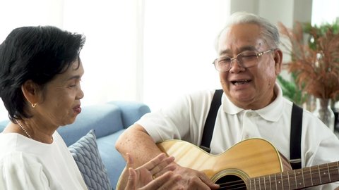 Asian senior couple sitting on the sofa and playing acoustic guitar together. Happy smiling elderly woman clapping hands while old 70s guitarist husband singing. Enjoying retirement life at home