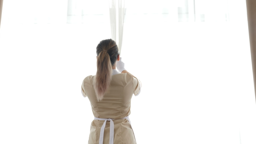 Back view of chambermaid in hotel opening curtains, going to clean up apartment. Cleaning in hotel. Housemaid in uniform opening curtain lace, letting light and fresh air into hotel room. 4 k footage Royalty-Free Stock Footage #1055933303