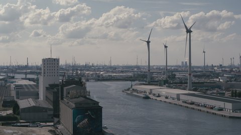 Aerial of the port of Antwerp depicting main canal old industrial buildings in the foreground and petrochemical industry in the background. Wind turbines spinning on a sunny day with some minor clouds