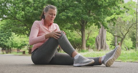 Senior fit Caucasian woman working out in the park wearing sports clothes, holding her injured knee with earphones on in slow motion. Retirement healthy lifestyle activity.