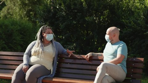 Pandemic concept. Two happy friends in medical mask keep social distance sitting and talking outside on bench in park at summertime. Couple prevent themselves from coronavirus covid spread.