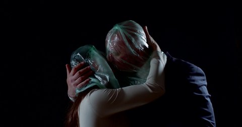 a man and a woman dressed in business clothes and standing on a black background. they kiss through plastic bags wrapped around their heads, then tear them apart