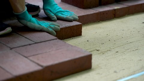 Low angle closeup of person installing brick pavers onto sand base wearing blue gloves.