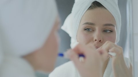 Young Caucasian woman in towel on head holding toothbrush brushing teeth looking in mirror.  Cleaning mouth doing morning oral dental care routine in bathroom.
