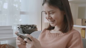 handle shot of excited young Asian woman playing games on smartphone in the living room at home.