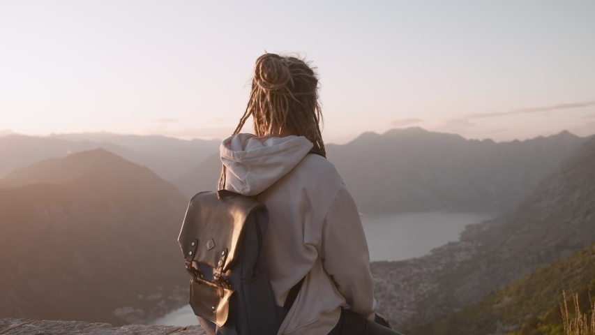 Back view of millennial dreadlocks woman  looking at nature background and raising hands, feeling free on top of the mountain cliff at sunset  in Montenegro. 
 | Shutterstock HD Video #1055939165